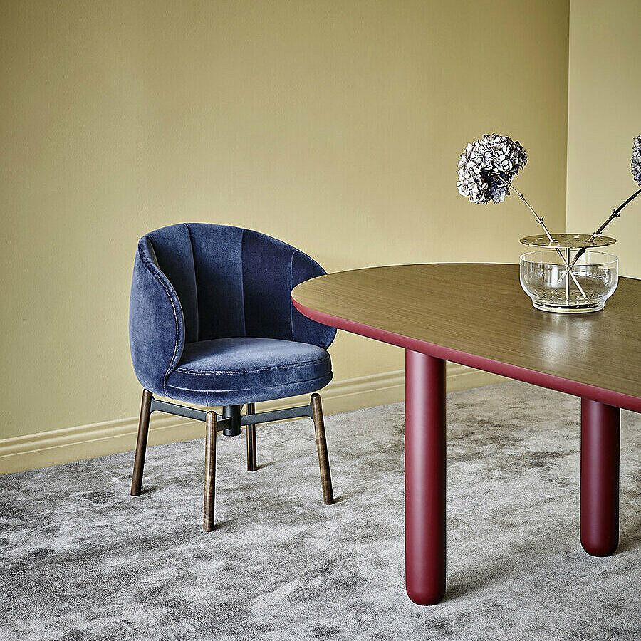 Vuelta FD Chair with dark blue velvet upholstery and swivel base with wooden legs in front of Vuelta FD Table with red legs