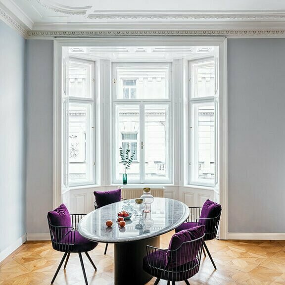 Esstisch mit grauer Marmorplatte und vier Stühlen mit Metallgestell und lila Sitzkissen vor großem Erkerfenster