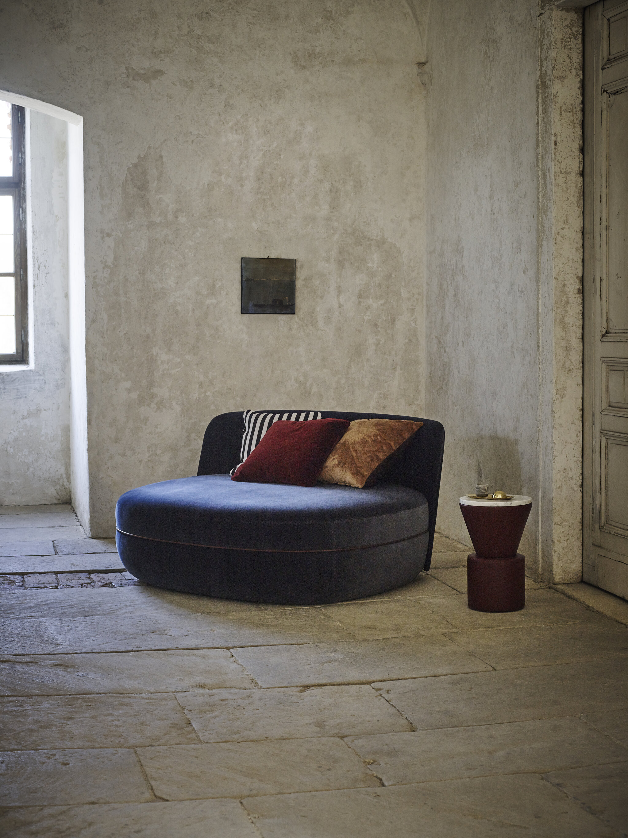 Miles Pouflounge in a blue velvet fabric and brown piping, next to it a Leather Side Table with white marble top and dark red base