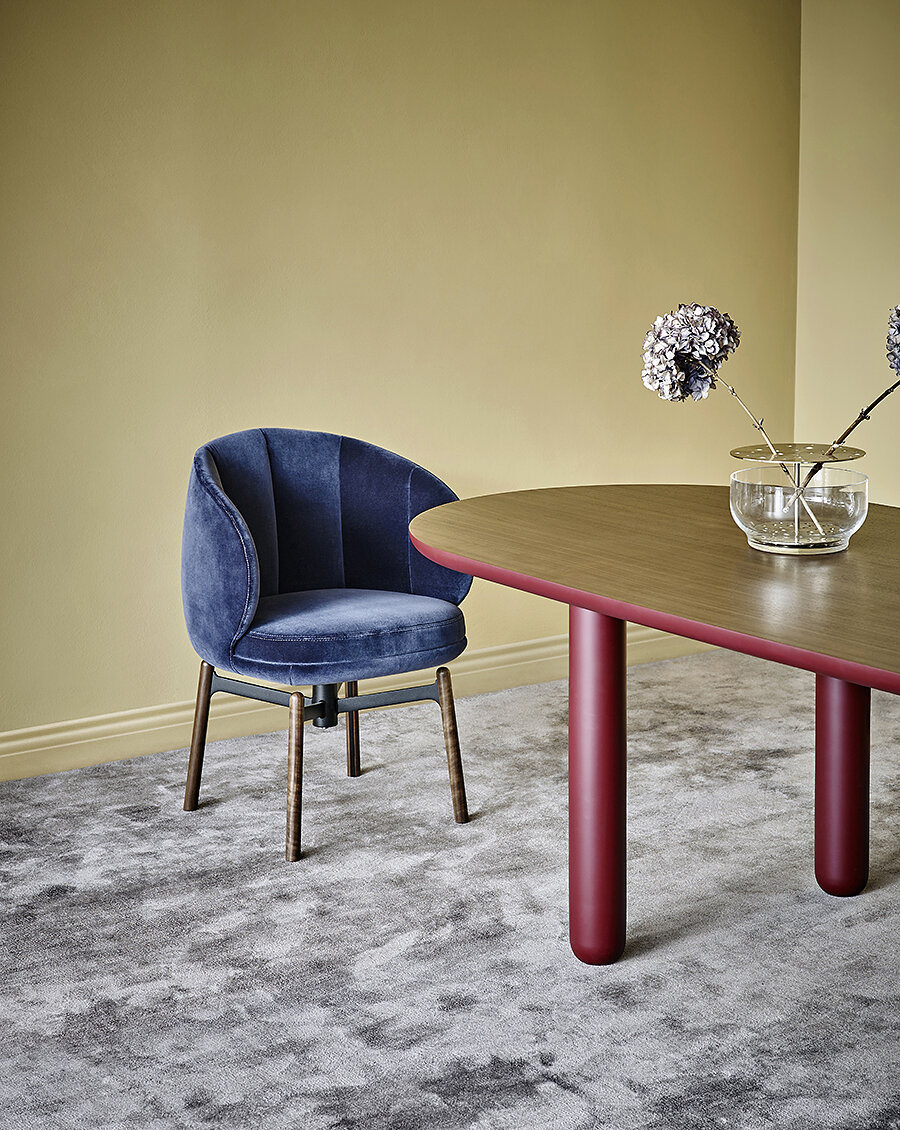 Vuelta FD Chair with dark blue velvet upholstery and swivel base with wooden legs in front of Vuelta FD Table with red legs